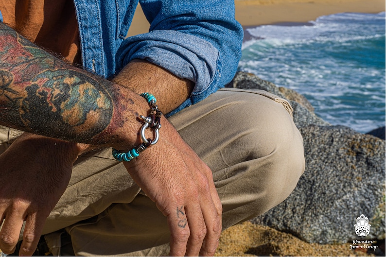 Op de afbeelding: Een turquoise en bruine kralen armband met een zilveren sluiting. De armband wordt gedragen op de pols van een persoon.