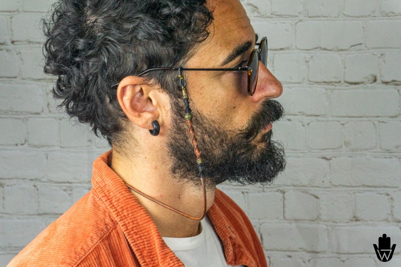 May include: A man wearing sunglasses with a brown leather cord attached to the frames. The cord has beads and metal accents.