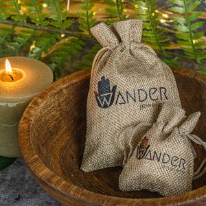 K&ouml;nnte beinhalten: Zwei kleine Jutes&auml;cke mit dem Aufdruck "Wander Jewelry". Die S&auml;cke liegen in einer Holzschale, im Hintergrund befindet sich eine brennende Kerze.