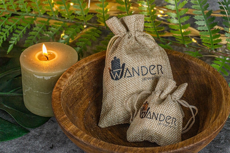 K&ouml;nnte beinhalten: Zwei kleine Jutes&auml;cke mit dem Aufdruck "Wander Jewelry". Die S&auml;cke liegen in einer Holzschale, im Hintergrund befindet sich eine brennende Kerze.