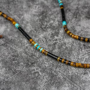 K&ouml;nnte beinhalten: Eine Perlenkette mit schwarzen, braunen und blauen Perlen. Die Kette ist aus nat&uuml;rlichen Steinen gefertigt und hat ein schlichtes Design.