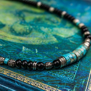 May include: A beaded necklace with black onyx and turquoise stones, accented with silver spacers. The necklace is lying on a blue and green patterned surface.