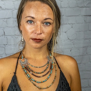 May include: A woman wearing a layered necklace with turquoise and brown beads and silver accents. The necklace has a crescent moon pendant. The woman is wearing a black top and has long brown hair.