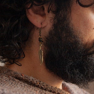 May include: A man with a beard wearing a gold feather earring with a black bead. The earring is hanging from his left ear.