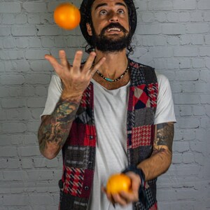 May include: A man with a beard and a hat is juggling three oranges in front of a white brick wall.