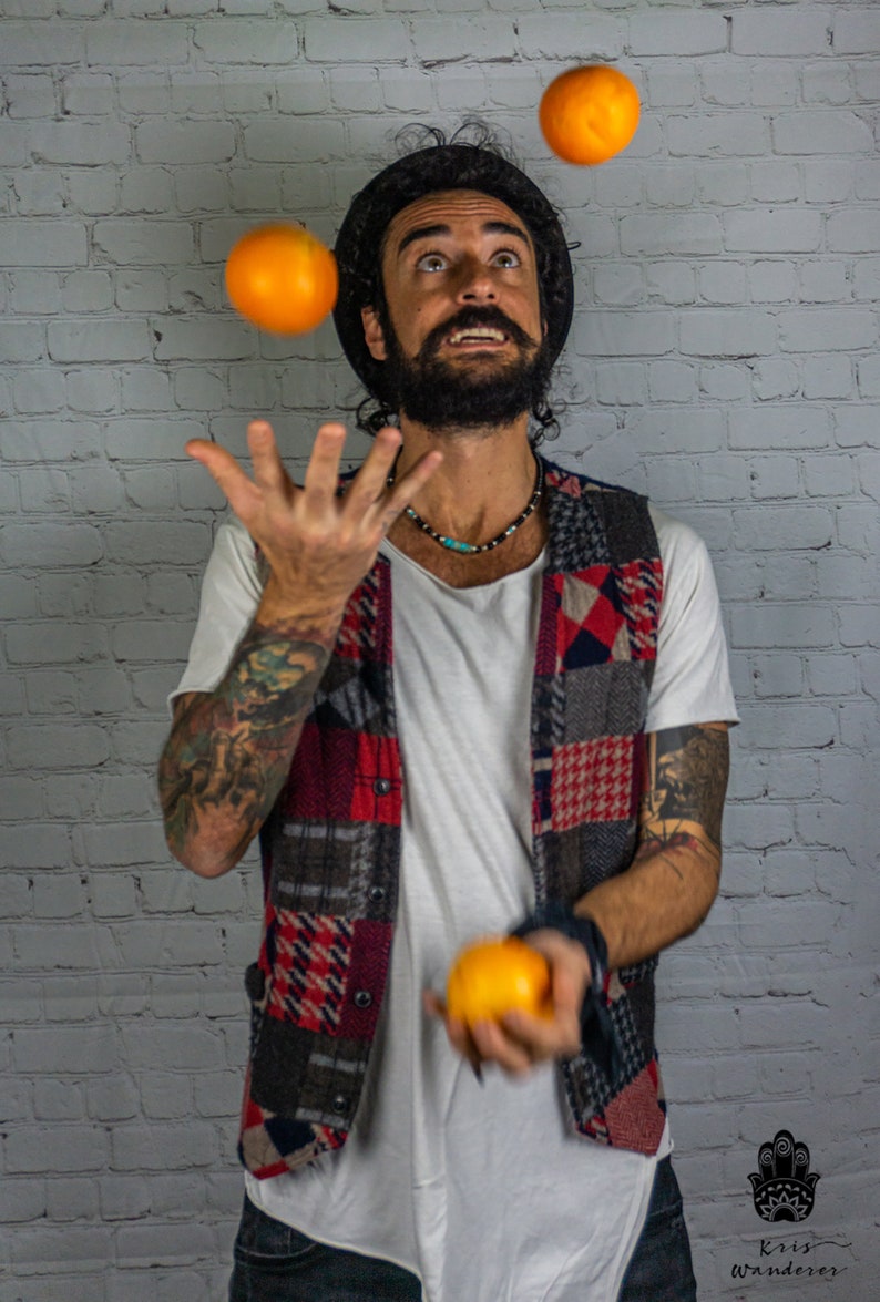 May include: A man with a beard and a hat is juggling three oranges in front of a white brick wall.