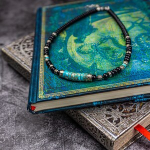 May include: A black and turquoise beaded necklace with silver accents. The necklace is lying on a blue and green book with a gold trim.