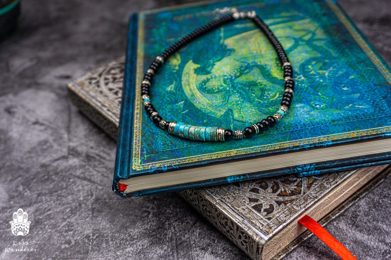 May include: A black and turquoise beaded necklace with silver accents. The necklace is lying on a blue and green book with a gold trim.