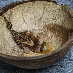 May include: A handcrafted earring featuring a brown and white feather, accented with bronze-colored beads and a small coin charm. The earring rests inside a textured, natural-toned coconut shell, set against a gray surface.