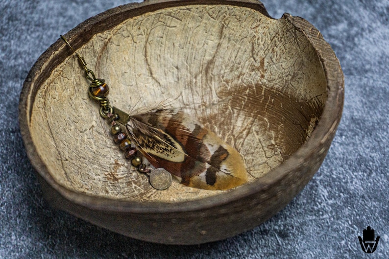 May include: A handcrafted earring featuring a brown and white feather, accented with bronze-colored beads and a small coin charm. The earring rests inside a textured, natural-toned coconut shell, set against a gray surface.