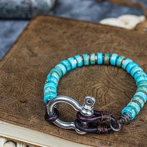 Op de afbeelding: Een turquoise edelsteen armband met een zilveren sluiting en bruine lederen accenten. De armband ligt op een bruin lederen oppervlak.