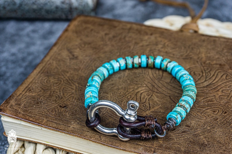 Op de afbeelding: Een turquoise edelsteen armband met een zilveren sluiting en bruine lederen accenten. De armband ligt op een bruin lederen oppervlak.
