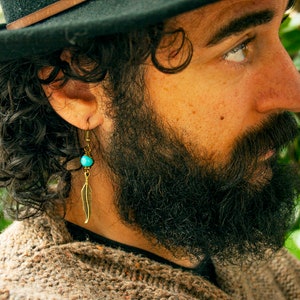 May include: A man with long curly dark hair and a beard is wearing a brown sweater and a black fedora. He is wearing a gold earring with a turquoise bead and a feather charm.