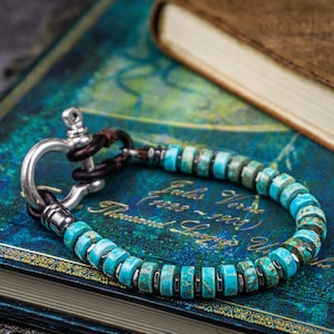 Op de afbeelding: Een turquoise en zilveren armband met een nautische sluiting. De armband is gemaakt van kleine, ronde turquoise kralen en een zilveren haaksluiting. De armband ligt op een blauw en gouden boek.