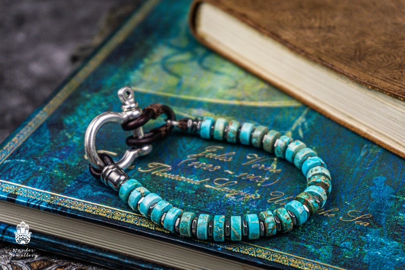 Op de afbeelding: Een turquoise en zilveren armband met een nautische sluiting. De armband is gemaakt van kleine, ronde turquoise kralen en een zilveren haaksluiting. De armband ligt op een blauw en gouden boek.