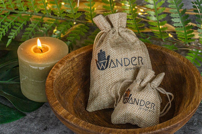 May include: Two burlap drawstring bags with the "Wander Jewels" logo sit in a wooden bowl. A lit candle and green foliage are in the background. The bags are different sizes and have a natural, rustic appearance.