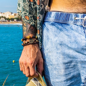 Könnte beinhalten: Ein Arm mit mehreren Perlenarmbändern in Braun- und Goldtönen. Die Person trägt hellblaue Jeansshorts und ein gemustertes grün-beiges Hemd. Der Hintergrund zeigt einen Strand und das Meer.