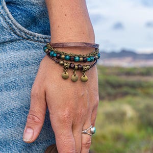 Peut inclure: Un bracelet multi-rangs composé d'un cordon en cuir plat marron, d'une fine chaîne en métal couleur bronze et d'un rang de perles alternant le marron foncé et le bleu sarcelle. Il comporte trois petites breloques rondes en métal bronze.