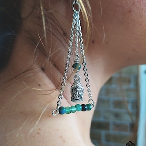 Può includere: Orecchini pendenti in argento con un ciondolo a forma di Buddha e perline verdi. Gli orecchini sono appesi all'orecchio di una persona.