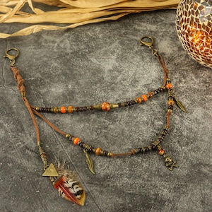 Könnte beinhalten: Eine braune Lederkordelkette mit orangefarbenen und braunen Perlen, einem Federschmuck und einem Totenkopf-Anhänger.