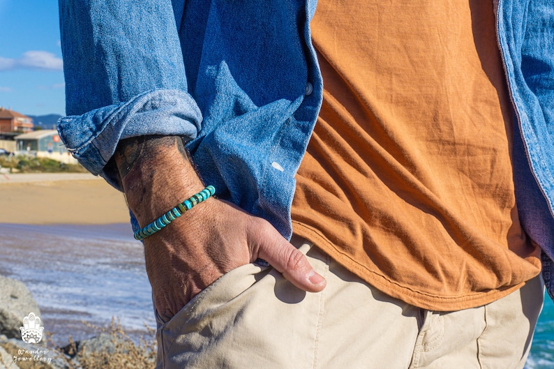 Op de afbeelding: Een man draagt een blauw denim shirt, een oranje t-shirt en een beige broek. Hij draagt een turquoise en bruine kralen armband om zijn linker pols.