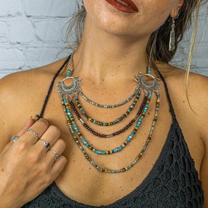 May include: A layered necklace with turquoise and brown beads and silver crescent moon pendants. The necklace is worn by a person with a white brick wall in the background.