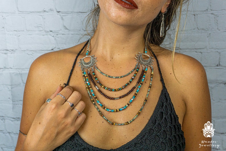 May include: A layered necklace with turquoise and brown beads and silver crescent moon pendants. The necklace is worn by a person with a white brick wall in the background.