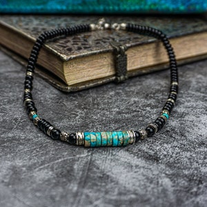 May include: A black beaded necklace with a turquoise and silver centerpiece. The necklace is lying on a gray textured surface.