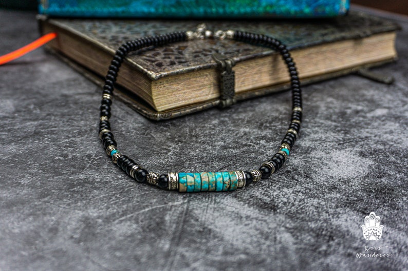 May include: A black beaded necklace with a turquoise and silver centerpiece. The necklace is lying on a gray textured surface.