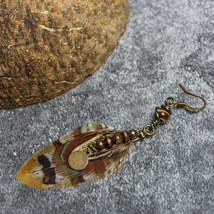 May include: A single feather earring with a bronze-colored hook. The feather is brown, tan, and black with a small, round, gold-colored accent. The earring also features small brown beads and a decorative bronze-colored metal piece.