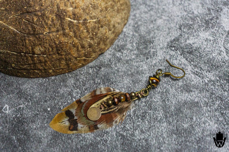 May include: A single feather earring with a bronze-colored hook. The feather is brown, tan, and black with a small, round, gold-colored accent. The earring also features small brown beads and a decorative bronze-colored metal piece.