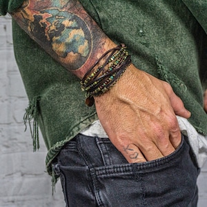 May include: A multi-strand leather bracelet with green and gold beads and a circular charm. The bracelet is worn on a wrist with a tattoo of a world map. The person is wearing a green jacket and dark jeans.
