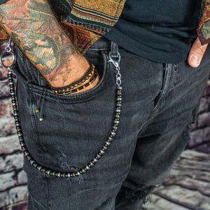 Può includere: Una catena per portafoglio con perline con chiusure color argento e un anello grande. La catena è attaccata a un paio di jeans neri strappati. Un braccialetto di perline abbinato è visibile nella tasca. Lo sfondo è un muro di mattoni.