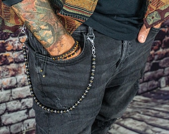 May include: A beaded wallet chain with silver-tone clasps and a large ring. The chain is attached to a pair of black distressed jeans. A matching beaded bracelet is visible in the pocket. The background is a brick wall.