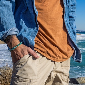 Op de afbeelding: Een man draagt een blauw denim shirt over een oranje t-shirt en een beige cargo broek. Hij draagt een turquoise kralen armband om zijn linker pols.