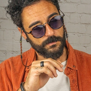 May include: A man with curly brown hair and a beard is wearing round tortoiseshell sunglasses with a beaded chain. He is wearing a rust-coloured corduroy shirt and a black ring on his finger.