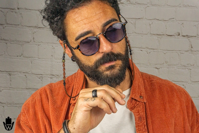 May include: A man with curly brown hair and a beard is wearing round tortoiseshell sunglasses with a beaded chain. He is wearing a rust-coloured corduroy shirt and a black ring on his finger.