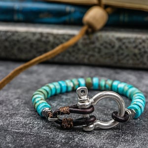 Op de afbeelding: Een turquoise en zilveren armband met een leren sluiting. De armband is gemaakt van kleine, ronde kralen en heeft een zilveren togglesluiting.