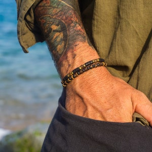 Pode incluir: Duas pulseiras de contas com detalhes em preto e dourado. As pulseiras são usadas no pulso, com o fundo de uma pessoa com uma camisa verde e uma tatuagem no braço. As pulseiras são feitas de pequenas contas redondas.
