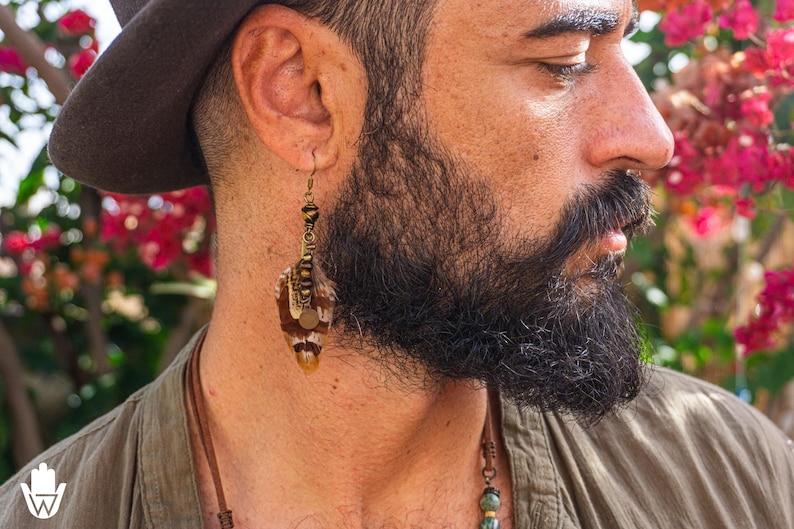 May include: A close-up of a person wearing a brown hat and a long, dangling earring. The earring features a combination of beads and feathers in shades of brown, tan, and gold. The person has a full, dark beard and is wearing a green shirt.
