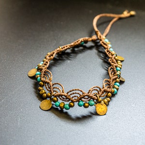 May include: A brown and turquoise beaded ankle bracelet with gold-toned metal accents. The bracelet is made with a macrame style knotting technique.