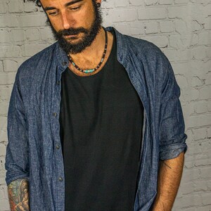 May include: A man wearing a black fedora, a black t-shirt, a blue denim shirt, and a beaded necklace with turquoise and black beads. He is standing in front of a white brick wall.