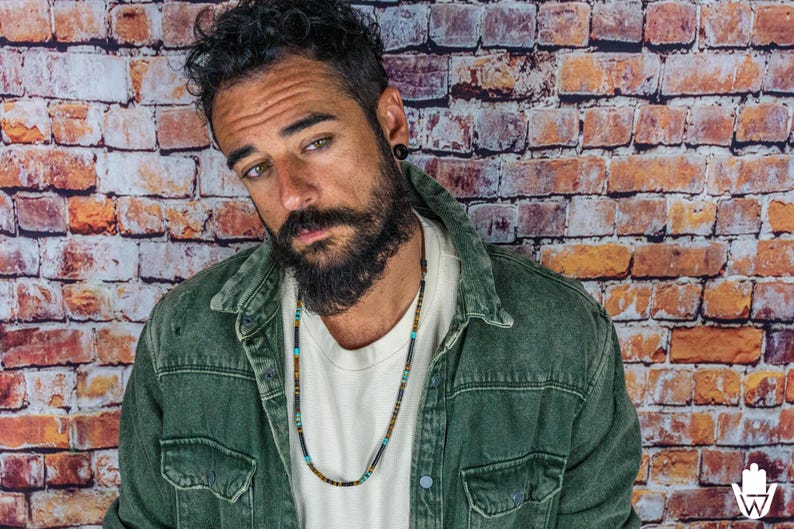 K&ouml;nnte beinhalten: Ein Mann mit Bart tr&auml;gt eine gr&uuml;ne Jeansjacke und ein wei&szlig;es T-Shirt mit einer mehrfarbigen Perlenkette.