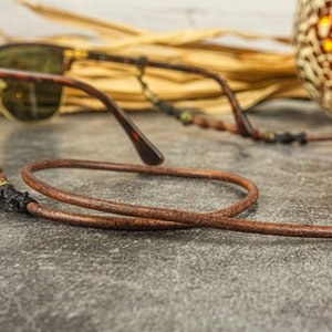May include: A brown leather eyeglass strap with a braided cord and metal accents. The strap is looped around a pair of brown sunglasses.