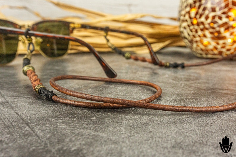 May include: A brown leather eyeglass strap with a braided cord and metal accents. The strap is looped around a pair of brown sunglasses.