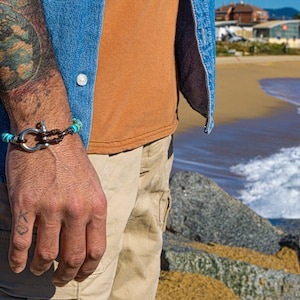 Op de afbeelding: Een bruin leren armband met turquoise kralen en een zilveren sluiting om de pols van een man. De armband zit om de pols van een man tegen een achtergrond van een strand en de oceaan.
