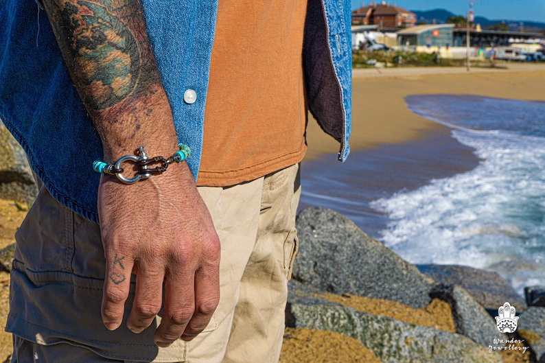 Op de afbeelding: Een bruin leren armband met turquoise kralen en een zilveren sluiting om de pols van een man. De armband zit om de pols van een man tegen een achtergrond van een strand en de oceaan.