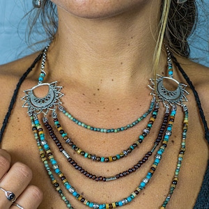 May include: Multi-layered necklace with turquoise, brown, and gold beads. The necklace features a silver pendant with a crescent moon design and a sunburst pattern. The necklace is worn by a person with a dark shirt.