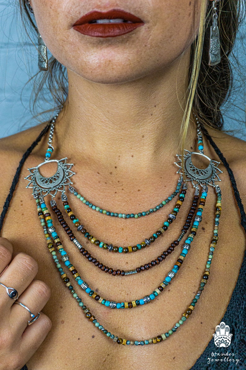 May include: Multi-layered necklace with turquoise, brown, and gold beads. The necklace features a silver pendant with a crescent moon design and a sunburst pattern. The necklace is worn by a person with a dark shirt.