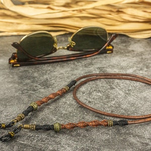 May include: A brown leather eyeglass strap with black and gold accents. The strap is twisted and has a decorative design.